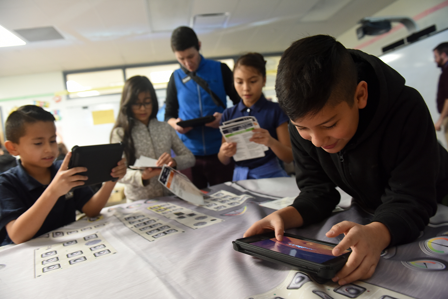 Several students lean on a table covered with a white cloth, engaging with printed materials and tablets